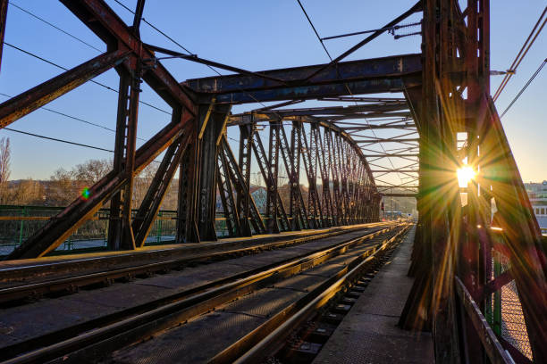 The setting sun casts golden light through the rusted steel truss frame of a railway bridge, creating a striking interplay of shadows and reflections. The converging lines of the tracks and beams draw the eye into the distance, evoking a sense of movement, history, and industrial beauty. The warm glow highlights the weathered textures of the aged metal, blending nature with engineering in a timeless composition.