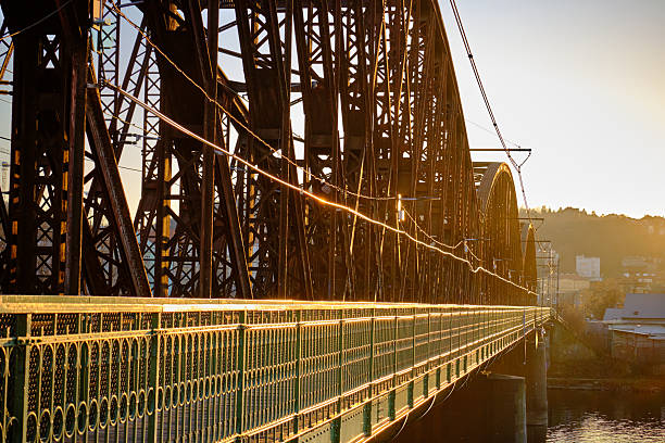 A steel truss bridge bathed in the golden light of the setting sun. The intricate lattice of riveted beams creates a rhythmic pattern, while the sun’s reflection on the railing adds a dynamic energy to the composition. The bridge’s repeating arches emphasize depth and perspective, showcasing the balance of strength and elegance in industrial-era engineering.