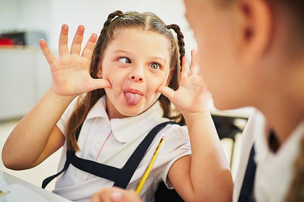 Elementary school girl playfully sticking out her tongue and making a funny face at her classmate during class, pulling her cheeks with her fingers and sharing a moment of joy