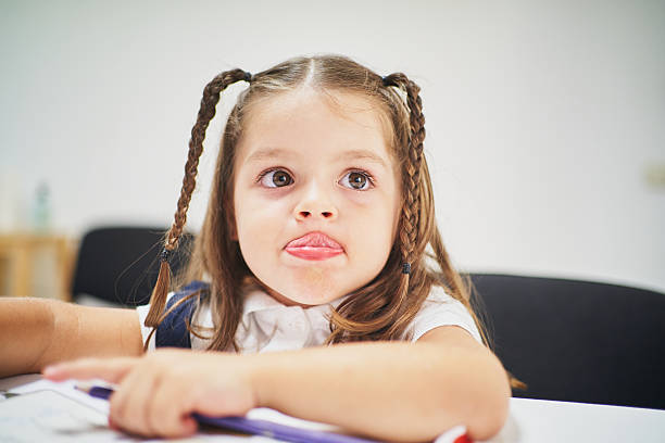 Preschool student sticking out her tongue while studying in a classroom, exuding a playful and carefree attitude towards learning, with braids framing her joyful expression