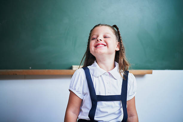 Portrait of a cheerful elementary school student smiling and looking up while standing in front of a chalkboard in a classroom, enjoying her school time