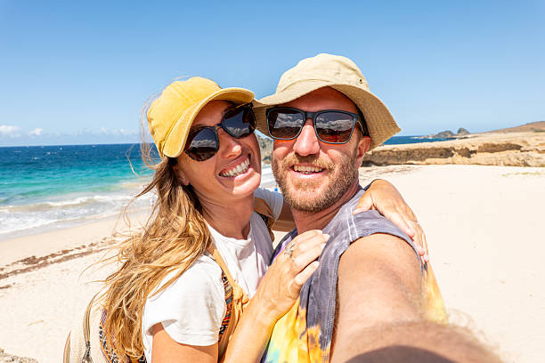 Smiling couple in hats and sunglasses taking a selfie on a sunny beach. Bright ocean and clear sky in the background. Perfect for travel and vacation themes.