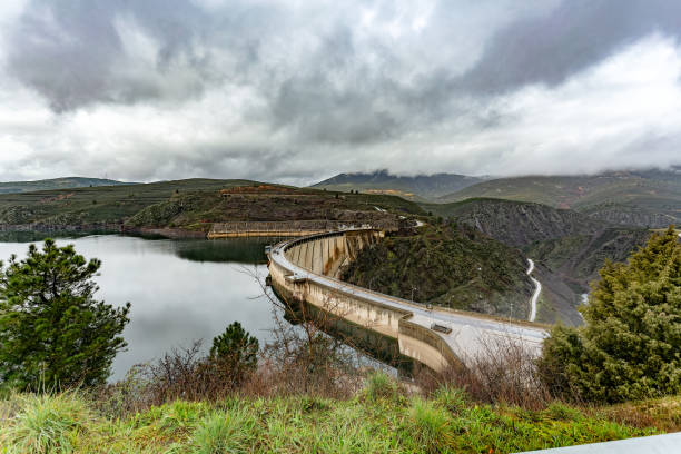 El Atazar reservoir, Madrid, Spain. A concrete dam curves gracefully through a mountain valley, creating a reservoir under a dramatic overcast sky