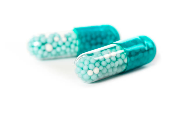 Two transparent pill capsules filled with blue and white beads are placed on a clean surface. The capsules are clearly visible, showcasing their contents in a clinical and sterile environment.