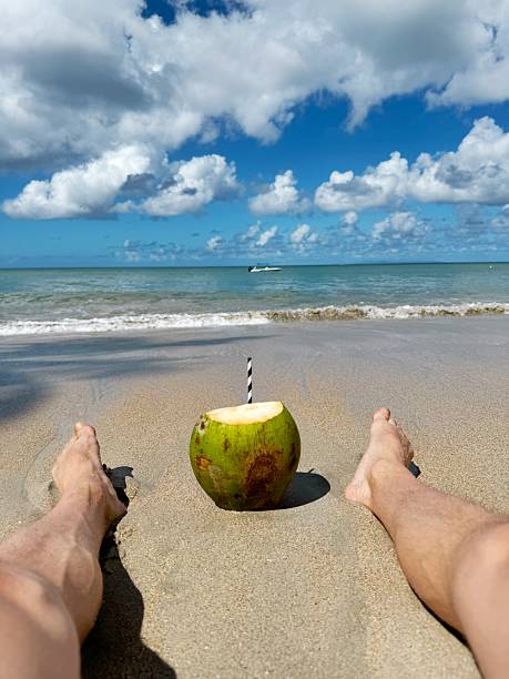 Person relaxing on a sandy tropical beach with a fresh coconut drink placed between their legs, facing the calm sea and vibrant sky filled with fluffy white clouds.