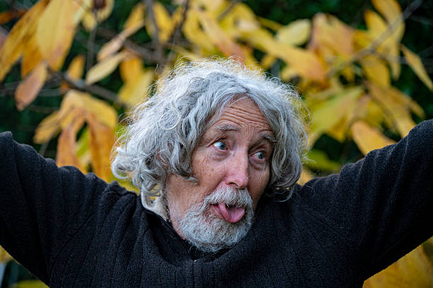 An elderly man with curly gray hair sticks out his tongue playfully, standing in front of vibrant autumn foliage, capturing a moment of lighthearted fun.