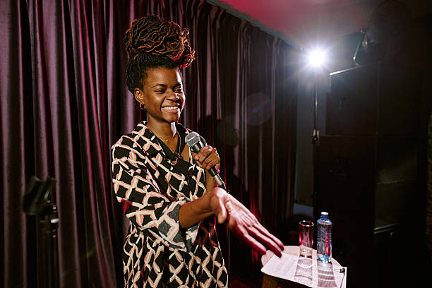 Smiling African American woman speaking on stage while holding microphone and extending hand. Engaging with audience in friendly and professional manner