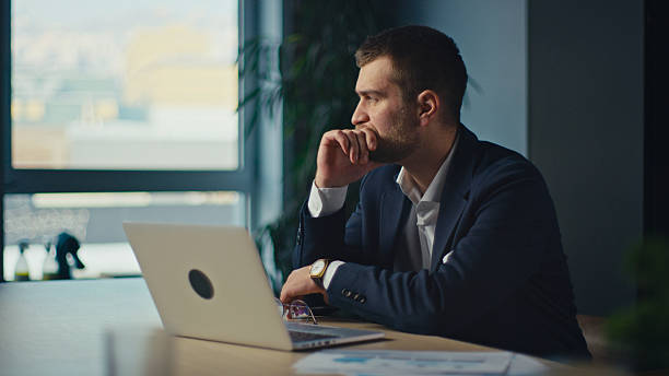 istock Young project manager or director in a suit sitting at a desk, deep in thought while looking at a laptop. Modern office setting. Concept of decision-making, corporate strategy, and business planning 2206682577