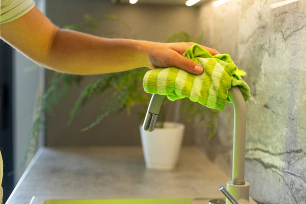 A close-up of a person's hand using a green cloth to clean a faucet, emphasizing cleanliness and a tidy, hygienic kitchen space.