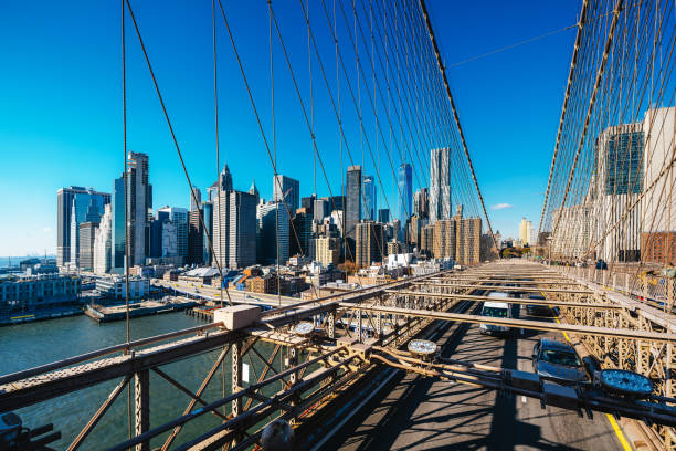View of the Brooklyn Bridge and downtown Manhattan skyline under a vibrant blue sky with sunlight highlighting the cityscape, creating a dynamic and iconic urban scene that represents New York City.