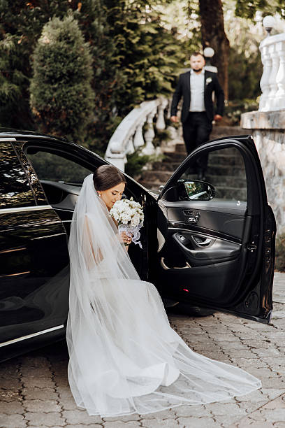 A bride is getting ready to get into a black car with a groom standing next to her. The car is parked on a driveway with a white railing in the background