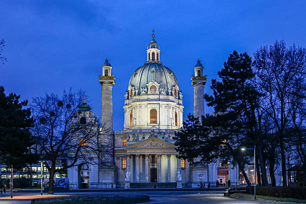 A large white building Karlskirche in Vienna Austria with a dome on top is lit up at night. The building is surrounded by trees and he is a church. The scene has a peaceful and serene mood