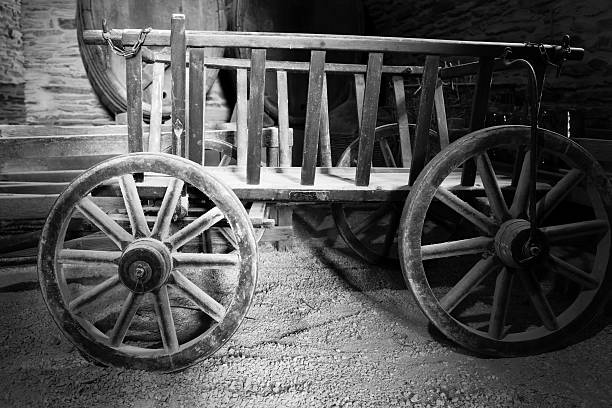 An aged wooden cart with spoked wheels stands prominently in a rustic barn. The dim light highlights its weathered detail, showcasing craftsmanship from another era.