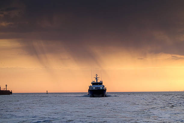 A departing vessel leaving Borkum harbor. The setting sun casts a warm glow across the water, illuminating a distant ship and the harbor entrance.  Sunrise with dramatic sky