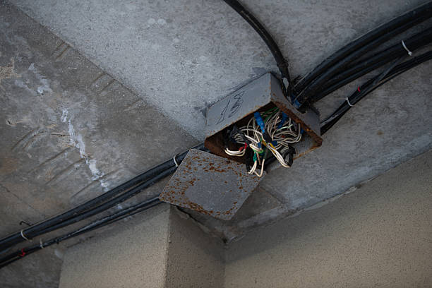 Rusty wiring box is mounted on the ceiling, revealing tangled cables in an incomplete construction site. The concrete surfaces show signs of wear and age, highlighting the building's unfinished state