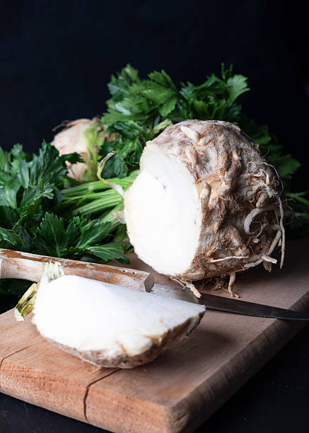 Celery root with rough, brown skin and white interior is partially sliced on a wooden board. Green leaves extend from the top, displaying rich foliage.