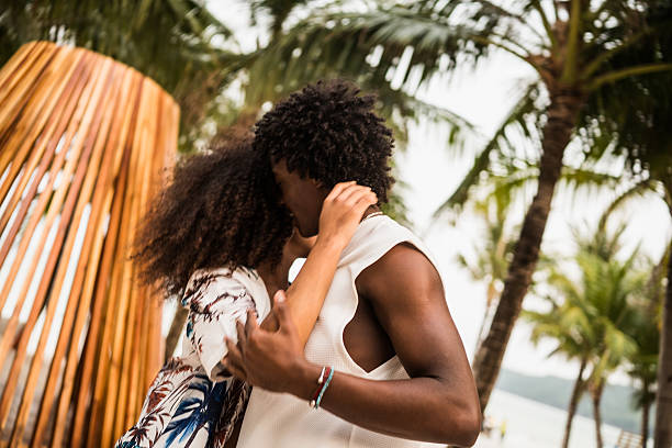 A joyful African American couple shares a warm hug on the beach, dressed in summer clothes, surrounded by palm trees and ocean views.