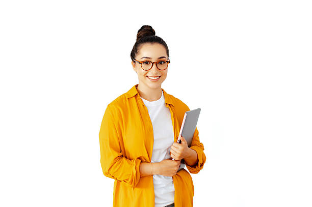 Pretty, positive caucasian brunette woman with glasses in a casual yellow shirt holds a tablet while standing against an isolated white background, she smiles confidently at the camera stock photo