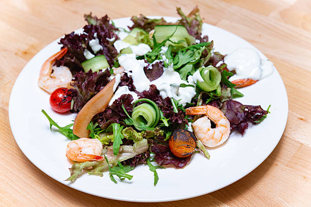 A vibrant shrimp salad with fresh greens, grilled cherry tomatoes, cucumber ribbons, poached pears, and creamy dressing, served on a white plate.