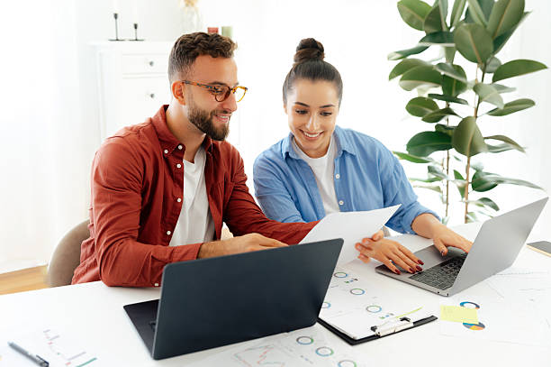 istock Two positive colleagues sit at their desks in an office, both working on laptops and smiling, they analyzing documents and showing them to the other, discussing work-related matters 2206756159