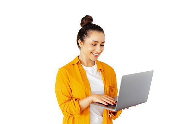 Happy caucasian woman in a casual yellow shirt types on a laptop while looking at the screen and smiles, she stands on isolated white background, symbolizing online work, and digital communication stock photo