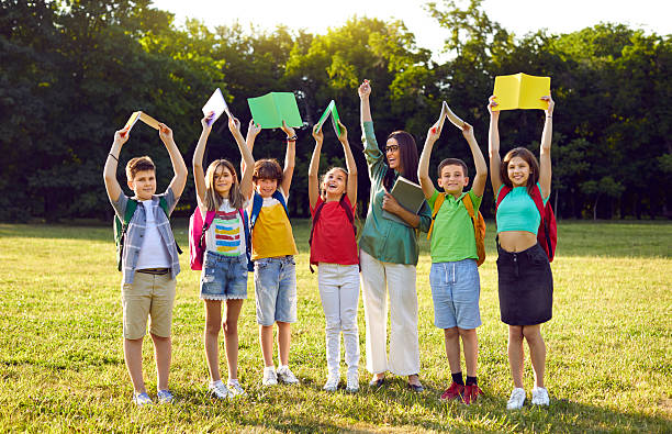 Female teacher and students with books having fun interesting class outside classroom. Portrait of happy, excited school teacher together with children standing in row in green field or schoolyard