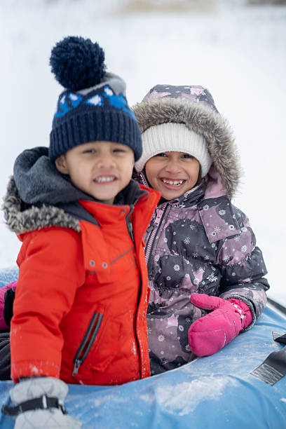 Two happy children dressed warmly in winter coats and hats, enjoying a fun day outdoors in a snowy environment. They are seated together, smiling and interacting in a friendly and playful manner.
