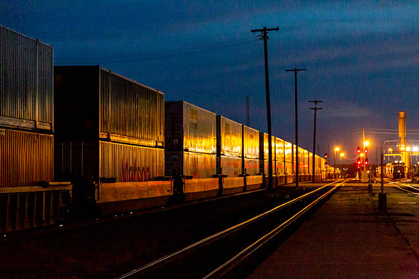 Newton KS, United States - June 18, 2018: A long and heavy freight train with double stack containers passes through the Kansas city of Newton at night, providing a 24/7 service to the railroad's customers. In the background, a service facility is providing round-the-clock maintenance for railroad rolling stock and equipment.