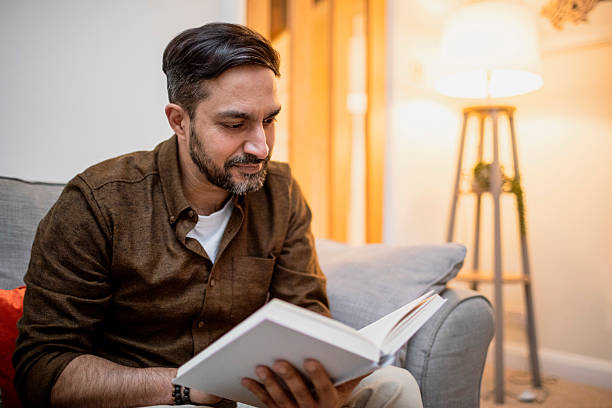 istock Man sitting on a sofa reading a book next to a floor lamp 2206794106