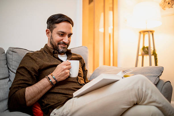 istock Man sitting on a sofa reading a book and holding a coffee mug 2206794141
