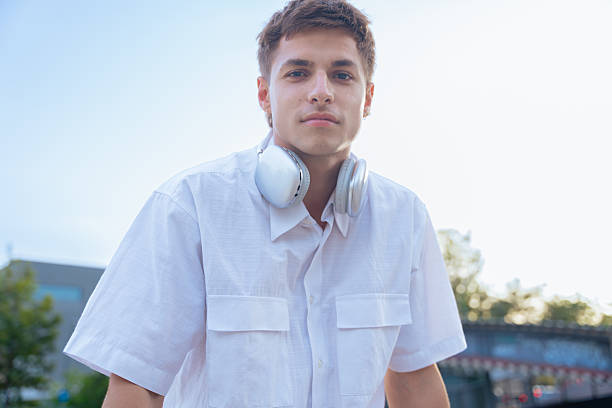 Young Caucasian man in white shirt wearing headphones around his neck outdoors. He looks confidently at the camera with a relaxed vibe, captured in a bright modern city atmosphere and sunlight.