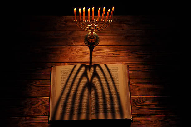A menorah illuminated by candles casts shadows over an open book on a wooden table, creating a warm, reflective ambiance.
