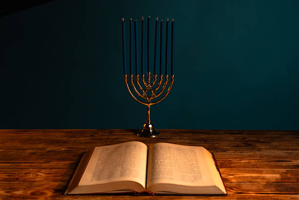A menorah with lit candles stands on a wooden table beside an open book, highlighting historical and cultural significance.