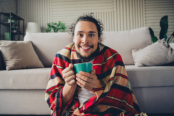 istock Young man relaxing at home with a cheerful expression, holding a warm drink wrapped in a cozy blanket 2206803561