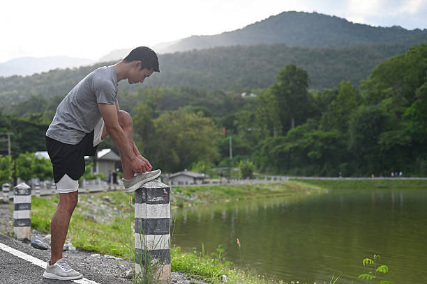 A young handsome man pauses by a lakeside, securing his shoelace on a pole, surrounded by lush greenery and serene waters