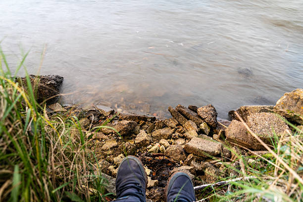 A person's feet rest on rocks by a calm lake, with water reflecting the overcast sky above.