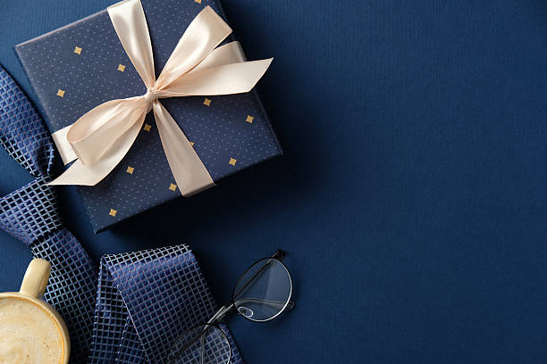 istock Gift box with beige ribbon, patterned blue tie, glasses, and coffee cup on a navy background. Father's Day, birthdays, or elegant celebration concept. 2206815614