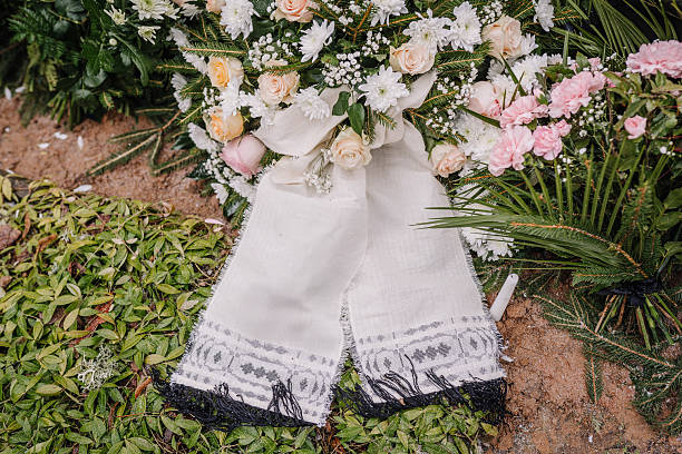 istock Funeral flower arrangement with white and pink blossoms, greenery, and a traditional patterned white cloth ribbon placed on a fresh grave. 2206819371