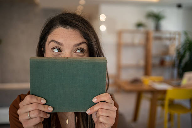 istock Playful woman hiding her face behind a book at home 2206845895