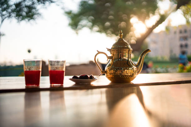 A golden teapot with two glasses of red tea and a plate of dates on a sunlit wooden table, surrounded by greenery in a serene Dubai cafe.