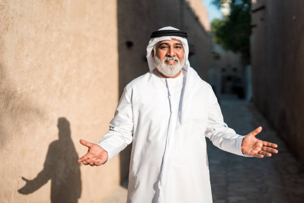An elderly Emirati man wearing traditional white attire and a headscarf stands outdoors in a bright, sunlit alley, smiling warmly with open arms, his shadow cast on the rustic walls around him.