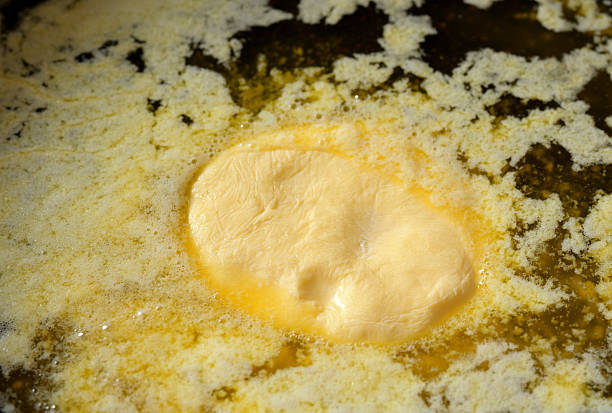 A close-up macro shot of artisanal butter melting in a hot pan. The golden-yellow butter forms a rich, creamy texture with foamy bubbles and a delicate milk skin on the surface. The high-resolution photograph captures the intricate details of the melting process, highlighting the fat separation and the transformation of the butter into clarified butter or ghee.
