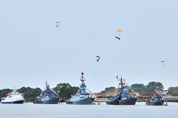 Airborne forces paratroopers flying Russian flag over naval forces parade warships along coastline, seafaring tradition of military ships formation at Navy Day, nautical spectacle of russian sea power