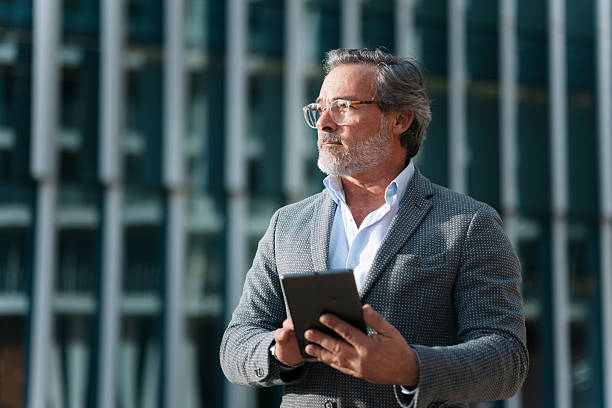 Confident businessman using digital tablet and looking away in front of modern office buildings