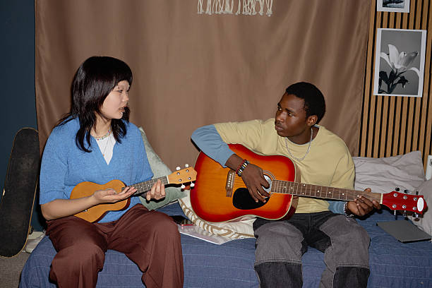 Asian woman and Black person sitting on sofa while playing guitar and ukulele. Relaxing atmosphere with framed picture and curtains in background
