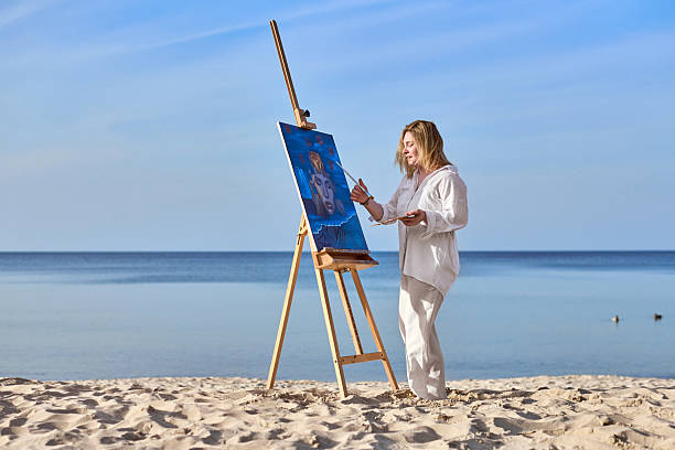 Young woman artist stands near ocean, painting on easel. Serene beach environment complements her creative process, evoking peace and inspiration