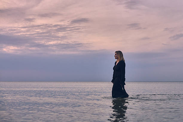 Young woman in black dress wades through calm ocean waters during tranquil sunset. Pink hues of sky evoke sense of solitude and peace, symbolizing introspection