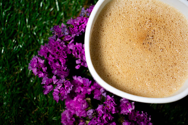 A close-up of a frothy cup of coffee on a yellow surface, next to vibrant purple flowers. The flowers have a delicate, papery texture and are clustered in small bunches, adding a vivid contrast against the smooth, dark liquid of the coffee. The bright yellow background enhances the colors, creating a cheerful and inviting scene.