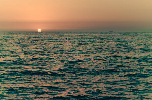 Tranquil ocean under orange-hued sky at dusk. Gentle waves reflect sunset light, creating serene atmosphere. Horizon showcases distant ship silhouette