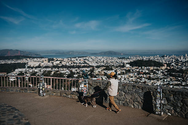 A Fillipino man in his 30s walks his pet dog at a public park overlooking San Francisco on a sunny and cool day, with the San Francisco Bay visible in the distance.